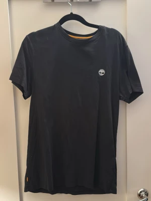 Svart Timberland t-shirt i bomull - Klassisk svart t-shirt från Timberland med liten vit logga på bröstet. T-shirten har rund halsringning och korta ärmar. Tillverkad i mjuk bomull för en skön och avslappnad stil. Perfekt till jeans eller shorts.