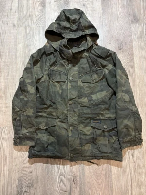 Abercrombie & Fitch  - Snygg camouflagefärgad vindjacka från Abercrombie & Fitch med huva och fyra framfickor med knappar. Jackan har en klassisk militärlook och är tillverkad i slitstarkt bomullsmaterial. Perfekt för dig som gillar streetwear och utilitystil. Passar M