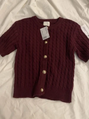 Vinröd stickad kofta från H&M - Säljer en vinröd kofta med korta ärmar och guldfärgade knappar framtill. Använd 1 gång. 