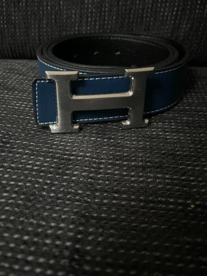 Blått skärp med H-spänne - Stilrent blått skärp med ett stort H-format spänne i silverfärg. Skärpet har vita sömmar längs kanterna. Perfekt accessoar för att lyfta din outfit. Ta insta box helst som frakt