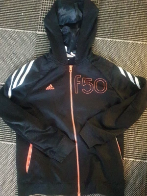 Svart Adidas hoodie med f50-tryck - Snygg svart hoodie från Adidas med orange dragkedja och f50-tryck på bröstet. Tre vita ränder på axlarna och orange detaljer vid fickorna. Klassisk huva och sportig look, perfekt för dig som gillar streetwear.