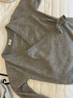 Grå omlottstickad tröja Kappahl - Mysig grå stickad tröja från Kappahl med omlottmodell och knytband i sidan. Tröjan har v-ringning och långa ärmar, perfekt för lager på lager. Materialet känns mjukt och bekvämt, och färgen är en klassisk ljusgrå ton.