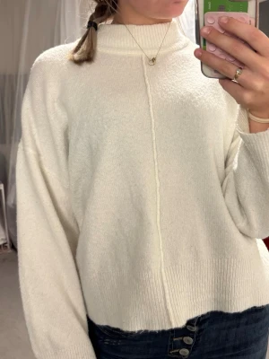 Vit stickad tröja - Säljer en vit stickad tröja med hög hals och synlig söm framtill. Tröjan har långa ärmar och ribbade muddar vid ärmslut och nederkant.