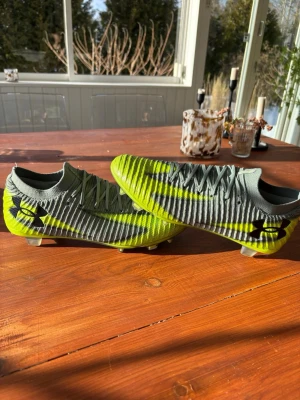 Under Armour Elite fotbollsskor - Snygga fotbollsskor från Under Armour i grått och neongrönt med svart logga på sidan. Skorna har en strumpliknande passform, ribbad ovandel och kolfiber sula för bra grepp och spänst på planen. Perfekta för dig som vill sticka ut på matchen! *Helt nya* storlek 42/2 men funkar för 41/2 och 42