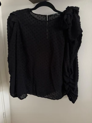 Svart blus med rosett från Zara - Snygg svart blus från Zara med diskret prickigt mönster i tyget. Blusen har lång ärm och en stor dekorativ rosett på ena axeln som ger en cool detalj. Tillverkad i ett tunt, transparent material som känns lätt och luftigt.