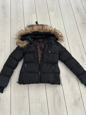 Svart pufferjacka från Hollies - Snygg svart pufferjacka från Hollies med luva och päls. Jackan har dragkedjor på fickorna och ett leopardmönstrat innerfoder som ger extra edge. Perfekt för kalla dagar när du vill hålla stilen. Nypris ca 5000-6000kr. Använd, pris går att diskutera. Frakt står köparen för