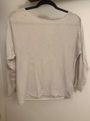 Beige tröja med rosett och öppning - Söt beige tröja med långa ärmar och en stor dekorativ rosett i nacken. Tröjan har en rundad halsringning och en öppen detalj i ryggen som ger en unik look. Perfekt för dig som gillar enkla men snygga detaljer.