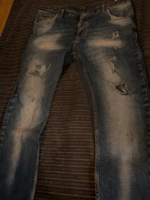 Blå slitna jeans med hål - Säljer ett par blå jeans med slitningar och hål på både lår och knän. Jeansen har en tvättad look med ljusare partier och detaljer som ger en riktigt trendig och avslappnad vibe. Klassisk femficksmodell och knappgylf. Perfekta för dig som gillar streetstyle.