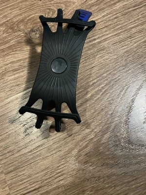 Svart cykelfäste för mobil - Flexibelt svart mobilfäste i silikon som enkelt monteras på cykelstyret. Perfekt för att hålla mobilen på plats under cykelturen och passar de flesta smartphones. Snygg design med solstrålemönster i mitten och elastiska hörn för säker fastsättning. Perfekt för sommar