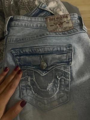 True Religion Joey ljusblå flare jeans - Säljer ett par ljusblå Joey low rise flare jeans från True Religion. Jeansen har en snygg fickdetalj med lock och knapp bak samt slitningar på fickan. Klassisk True Religion-logga på bakfickan och patch i midjan. Tillverkade i denim med en avslappnad passform.