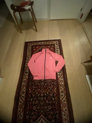 Rosa träningsjacka från Adidas - Snygg rosa träningsjacka från Adidas med svart dragkedja och vit logga på bröstet. Jackan har hög krage och långa ärmar, perfekt för träning eller sportiga dagar. Tillverkad i ett lätt och funktionellt material.