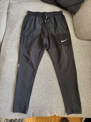 Svarta träningsbyxor running från Nike - Säljer ett par svarta träningsbyxor running från Nike med dragsko i midjan och vita loggor på benet. Byxorna har smal passform och är tillverkade i ett lätt och stretchigt material som passar perfekt för träning eller löpning.