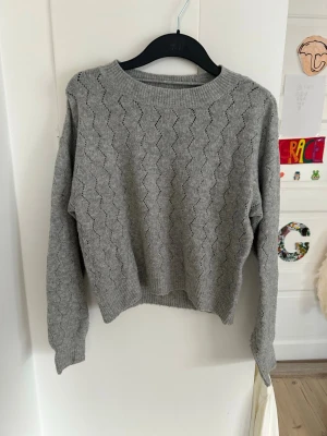 Grå stickad tröja med mönster vero moda - Mysig grå stickad tröja från Vero Modamed diskret zigzag-mönster över hela plagget. Tröjan har rund halsringning, långa ärmar och ribbade muddar vid ärmslut och nederkant. Perfekt för lager-på-lager och enkel att matcha med jeans eller kjol. Storlek S
