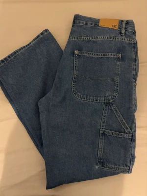 Blåa jeans från 157 med stora fickor - Säljer ett par blåa jeans från 157 med raka ben och stora, praktiska fickor på sidan. Jeansen har en klassisk arbetsbyx-look och är gjorda i slitstarkt denimtyg. Perfekta för dig som gillar en avslappnad och trendig stil.