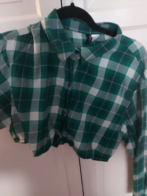Grön rutig cropped skjorta - Snygg grön och vit rutig cropped skjorta från H&M. Skjortan har klassisk krage, långa ärmar och knäppning framtill. Perfekt för dig som vill ha en avslappnad men trendig stil. Tillverkad i mjuk bomull som känns skön mot huden.