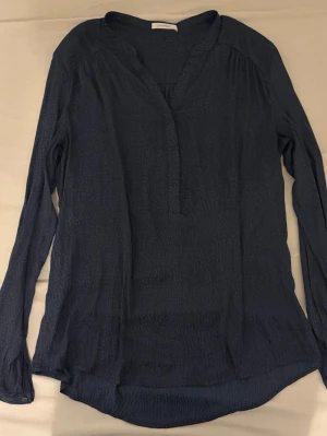 Mörkblå blus från Promod - Stilren mörkblå blus från Promod med diskret mönster och v-ringad hals. Blusen har långa ärmar och är tillverkad i ett lätt, luftigt tyg som ger en avslappnad men ändå chic look. Perfekt att matcha med jeans eller kjol.