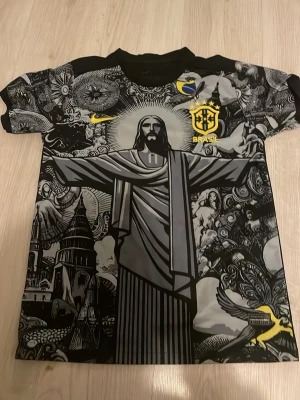 Brasil t-shirt med Kristusstatyn - Unik svart och grå Brasil t-shirt från Nike med stort tryck av Kristusstatyn i Rio och detaljerade grafiska mönster. Gula Brasil-loggan och Nike swoosh på bröstet. Perfekt för dig som gillar fotboll och streetwear.
