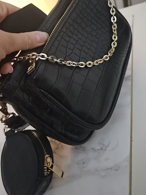 Svart croco handväska med kedja - Snygg svart handväska i croco-mönstrat konstläder med guldfärgade detaljer och kedja. Väskan har flera fack med dragkedja och en liten rund coin purse som hänger på sidan. Perfekt accessoar för att lyfta din outfit. Väldigt fint skick.