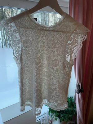 Vit spetsblus från Pieces - Säljer en superfin vit blus från Pieces med detaljerad spets och blommigt mönster. Blusen är ärmlös med vackra spetskanter vid axlarna och har en feminin, romantisk look. Perfekt att styla med jeans eller kjol för en trendig outfit.
