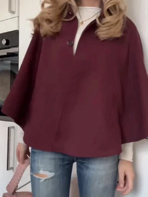 Vinröd cape-kappa med knapp - Stilren vinröd cape-kappa med vid passform och en dekorativ knapp framtill. Kappan har en hög krage och är tillverkad i ett mjukt tyg som ger ett elegant intryck. Perfekt för dig som vill ha en trendig och unik ytterjacka.