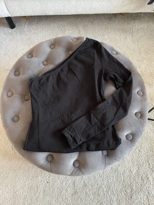 Svart topp - Snygg svart topp med en axel och lång ärm. Tight passform och stilren design från Nelly. Passar perfekt till vardags eller fest.