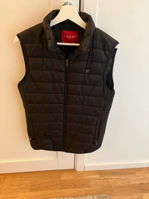 Svart dunväst från Guess - Snygg svart dunväst från Guess med hög krage och dragkedja framtill. Västen har två sidofickor med dragkedja och quiltad design. Perfekt för lager på lager och ger en clean look till din outfit.