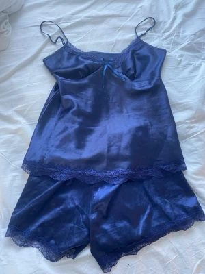 Mörkblå satinpyjamas med spets - Säljer ett mörkblått sovset i satin med linne och shorts. Setet har tunna axelband, spetsdetaljer vid kanterna och en liten rosett framtill på linnet. Materialet är glansigt och mjukt, perfekt för en lyxig känsla. 