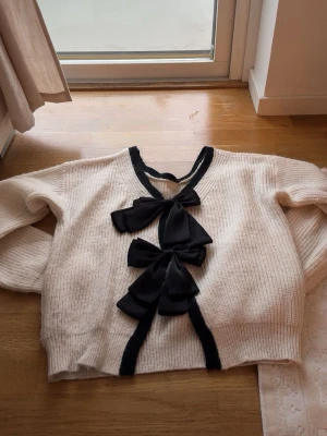 Vit stickad tröja med svarta rosetter - Trendig vit stickad tröja med v-ringning och tre stora svarta rosetter framtill. Svarta detaljer längs kanterna ger en snygg kontrast. Tröjan har lång ärm och är gjord i ett mjukt, ribbstickat material. Perfekt för dig som gillar statement-plagg. 