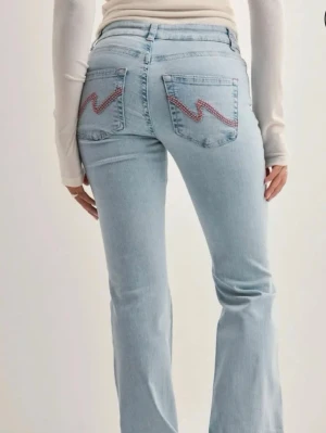 Nelly bootcut jeans -  Ljusblå bootcut jeans med slitna detaljer vid knäna och cool zigzag-broderi på bakfickorna. Jeansen har låg midja och är tillverkade i klassisk denim. Perfekta för en avslappnad och trendig look.