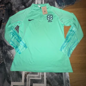 Nike Brasil långärmad fotbollströja S - Snygg mintgrön långärmad fotbollströja från Nike med Brasil-logga och dragkedja vid halsen. Ärmarna har ett coolt rutigt mönster i blå toner. Perfekt för träning eller när du vill representera Brasilien. Materialet är lätt och andas, vilket gör den skön att ha på sig.
