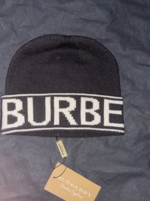 Svart mössa från Burberry - Snygg svart mössa från Burberry med vit logotyp runt hela kanten. Klassisk beanie-modell i mjukt stickat material. Perfekt accessoar för dig som vill ha en stilren och trendig look med tydlig branding.