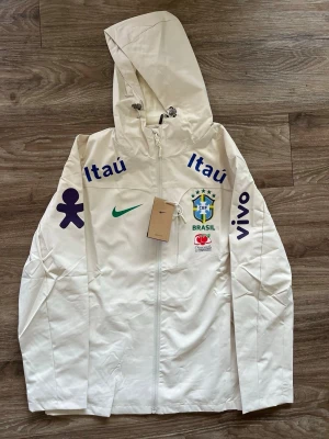 Vit Nike Brasil vindjacka med huva - Snygg vit vindjacka från Nike med Brasilien-logga och sponsormärken i blått och grönt. Jackan har huva med dragsko, hel dragkedja framtill och tryck på både bröst och ärmar. Perfekt för dig som gillar sportig stil och vill sticka ut med landslagskänsla.