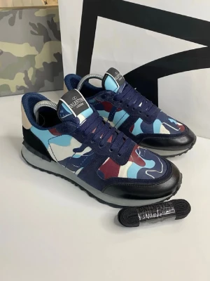 Valentino blå camo sneakers - Snygga sneakers från Valentino med blå, vita och röda camo-detaljer. Skorna har mörkblå snörning, svart sula och svarta läderdetaljer. Ovandelen är i textil och läder, med logga på plösen. Perfekta för dig som vill sticka ut med en cool streetstyle.