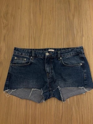 Mörkblå jeansshorts med rå kant - Säljer ett par mörkblå jeansshorts från Nelly med rå, fransig kant och klassisk femficksdesign. Shortsen har låg midja och är gjorda i slitstark bomull. Perfekta för varma dagar och en avslappnad stil.