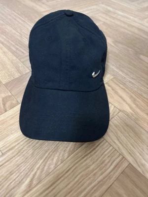 Svart Nike keps med liten Swoosh - Stilren svart keps från Nike med den klassiska lilla Swoosh-loggan i silver på framsidan. Kepsen har böjd skärm och justerbar rem bak. Tillverkad i mjukt bomullstyg som ger en avslappnad och sportig look.