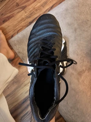 Nike svart-vita fotbollsskor med dobbar - Säljer ett par svarta fotbollsskor från Nike med vita detaljer och metall-dobbar för bästa grepp på planen. Skorna har snörning och en slitstark ovandel i syntetmaterial. Perfekta för matcher på naturgräs där du vill ha stabilitet och kontroll.