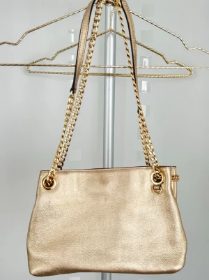 Guldig handväska från Michael Kors - Snygg guldig handväska från Michael Kors i metallicläder med kedjehandtag i guldfärg. Bra accessoar för dig som vill sticka ut. Använt en ett par gånger. 