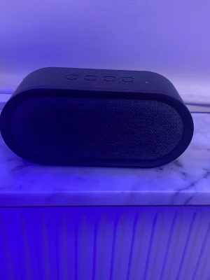 Svart bärbar Bluetooth-högtalare - Snygg svart bärbar Bluetooth-högtalare med rundade kanter och tygfront. Högtalaren har fyra knappar på ovansidan för enkel styrning av musik och volym. Perfekt för att ta med sig överallt och spela upp favoritlåtarna trådlöst.