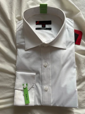 Vit slim fit skjorta från Dressmann - Klassisk vit skjorta från Dressmann i slim fit-modell. Skjortan har lång ärm, knappar framtill och vid manschetten samt en stilren krage. Perfekt för en clean och snygg look.