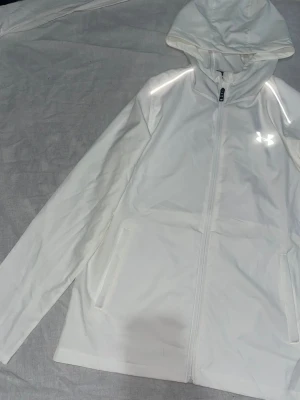 Vit hoodie från Under Armour - Snygg vit hoodie från Under Armour med dragkedja framtill och reflekterande logga på bröstet. Tillverkad i ett lätt och funktionellt material som andas, perfekt för träning eller chill. Utrustad med huva och två fickor på sidorna.