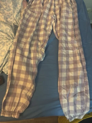 Pjamas sätt - Mysiga pyjamasbyxor med lila och vitt rutmönster. Byxorna har resår i midjan och vid bensluten för extra komfort. Perfekta för slappa dagar hemma eller en skön natts sömn. Tillverkade i mjukt tyg som känns skönt mot huden. Pjamassätt