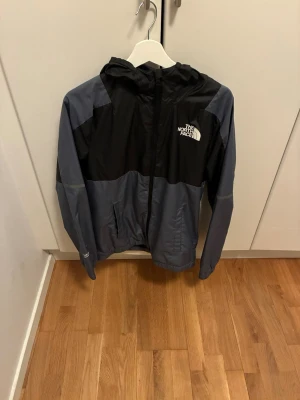 Svart och grå vindjacka The North Face - Snygg vindjacka från The North Face i svart och mörkgrått. Jackan har huva, dragkedja framtill och två sidofickor. Perfekt för blåsiga dagar och enkel att ta med sig. Logga på bröstet och lätt material som andas.