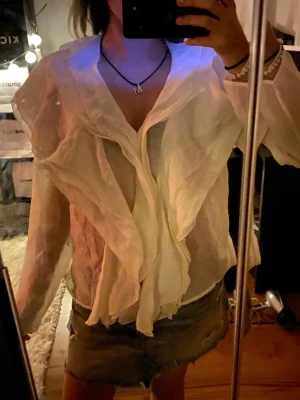 Vit volangblus med transparent tyg - Säljer en vit blus med långa ärmar och volangdetaljer framtill. Blusen är gjord i ett tunt, transparent material som ger en luftig känsla och har en djup v-ringning. Perfekt för dig som gillar statement-plagg och vill ha något extra till din outfit.