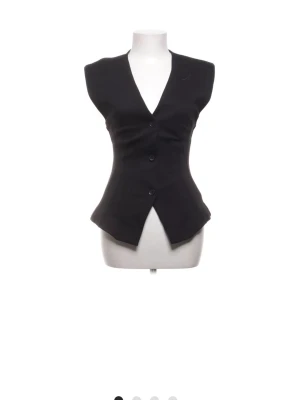 Svart v-ringad väst med knappar - Snygg svart väst med v-ringning och tre knappar framtill. Västen har en figurnära passform och markerad midja som ger en stilren siluett. Perfekt att styla över en skjorta eller t-shirt för en trendig look.