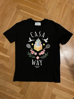 Svart t-shirt från Casablanca - Svart t-shirt från Casablanca med färgglatt tryck framtill som visar en byggnad, två duvor, tennisracketar, rosor och texten 'CASA WAY'. Klassisk passform och rund hals. Perfekt för dig som gillar unika prints och sportiga vibbar.