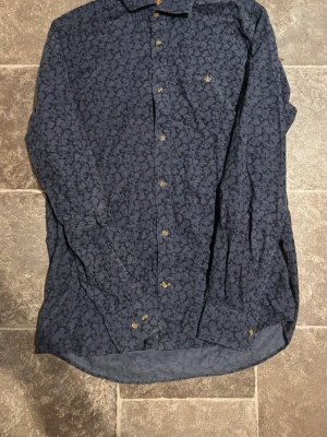 Mörkblå blommig skjorta med brodyr - Snygg mörkblå skjorta med blommigt mönster och guldfärgad broderad detalj på bröstet. Skjortan har långa ärmar och knappar framtill. Perfekt för dig som vill sticka ut lite extra.