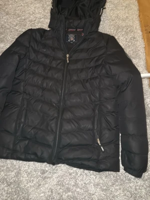 Svart pufferjacka Vinson Polo Club - Snygg svart pufferjacka från Vinson Polo Club med huva och dragkedja framtill. Jackan har två sidofickor med dragkedja och quiltad design som ger en sportig look. Perfekt för kalla vinterdagar och riktigt skön att bära. Kan va lite trög att stänga ibland 