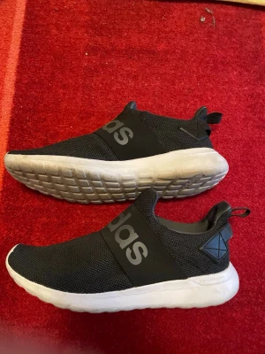 Svarta Adidas Cloudfoam slip-on sneakers - Säljer ett par svarta Adidas Cloudfoam slip-on sneakers med bred resår över foten och stor logga. Ovandelen är i mesh-tyg och sulan är vit och tjock för extra komfort. Snygg och enkel design med dragflik bak för lättare påtagning.