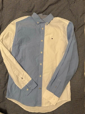 Blåvit skjorta från Tommy Hilfiger - Säljer en stilren skjorta från Tommy Hilfiger i blått och vitt colorblock-mönster. Skjortan har klassisk krage, långa ärmar och knappar framtill. Liten broderad logga på bröstet. Perfekt för dig som gillar en clean och modern look.