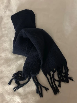 Mörkblå halsduk med fransar - En enkel och stilren mörkblå halsduk med fransar i båda ändar. Halsduken är mjuk och har en klassisk rektangulär form, perfekt att vira runt halsen för extra värme. Materialet känns fluffigt och bekvämt mot huden.